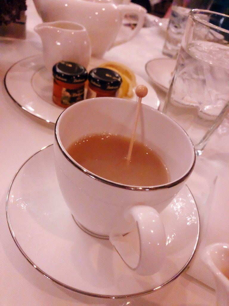 A white teacup filled with with warm gingerbread tea. In the background, there are a few miniature jars of honey, a mini jug of milk and a few lemons to add for flavor.